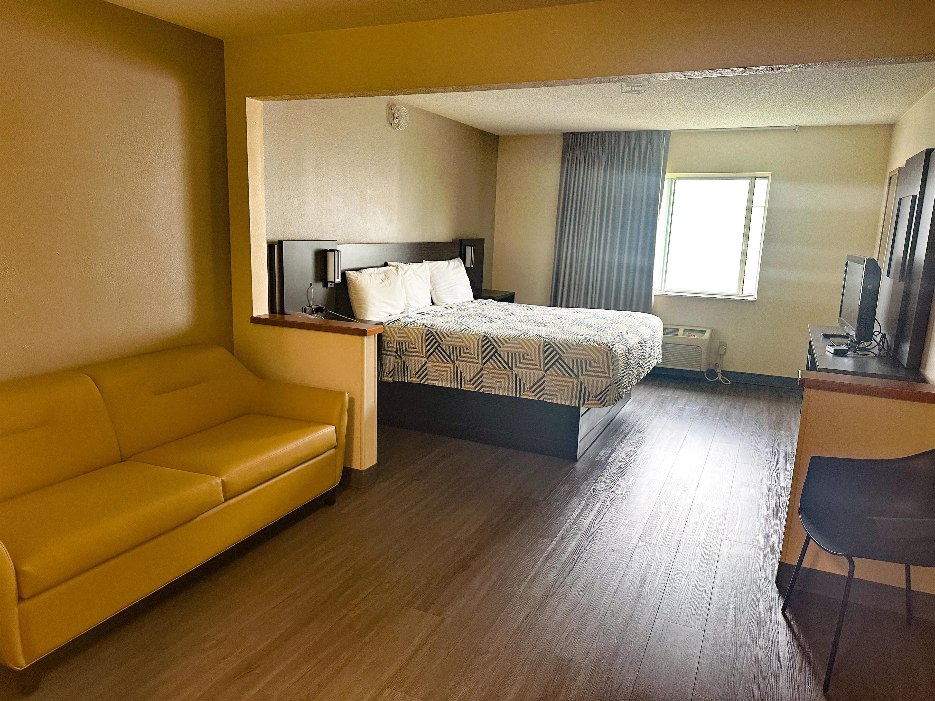 A hotel room with a bed, a yellow sofa, a desk with a computer, and a window with curtains.
