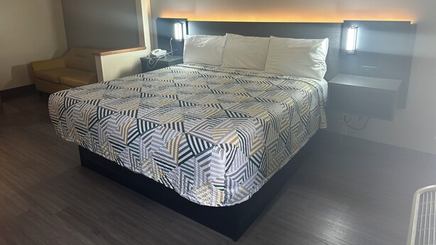 A hotel room with a large bed, a bedside table with a lamp, a chair, and a wall-mounted phone. - Motel 6 Anderson, IN - Indianapolis (Anderson)
