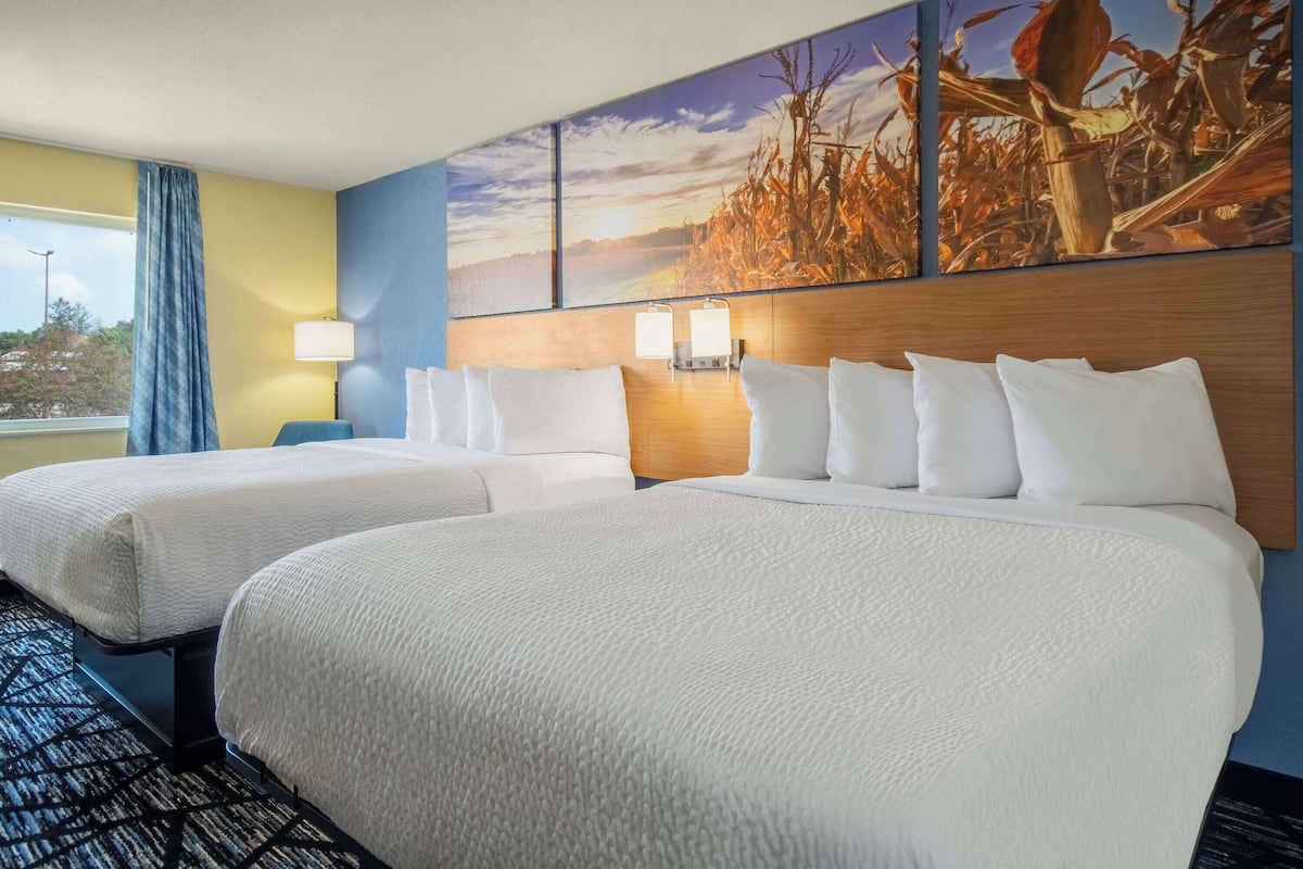 A hotel room with two beds, a large window with curtains, a wall-mounted lamp, and a painting of a landscape.