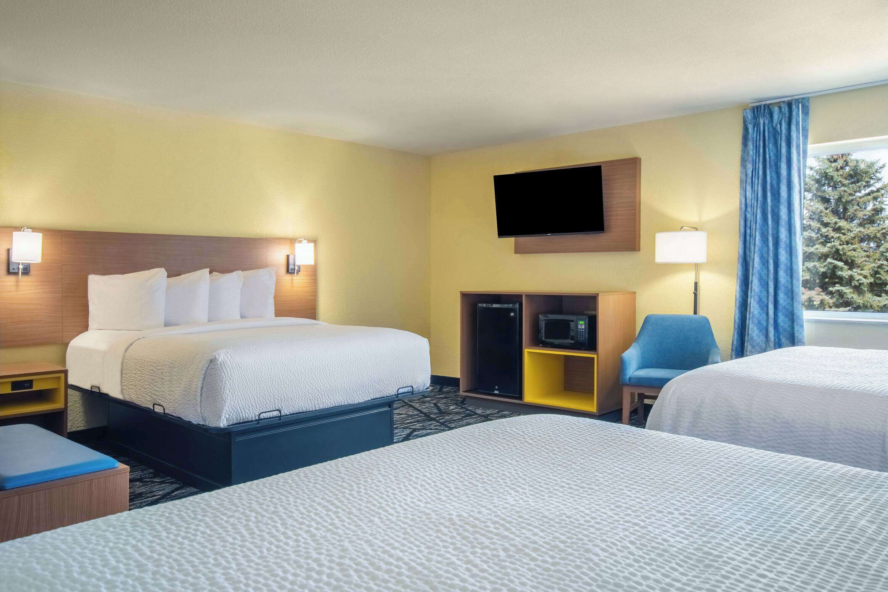 A hotel room with two beds, a flat-screen TV mounted on the wall, a desk with a lamp, and a view of trees through the window.