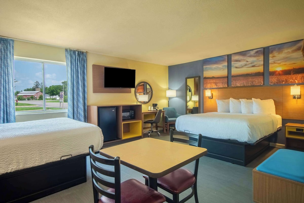 A hotel room with two beds, a desk, a chair, a TV, a desk lamp, a mirror, and a painting of a sunset.