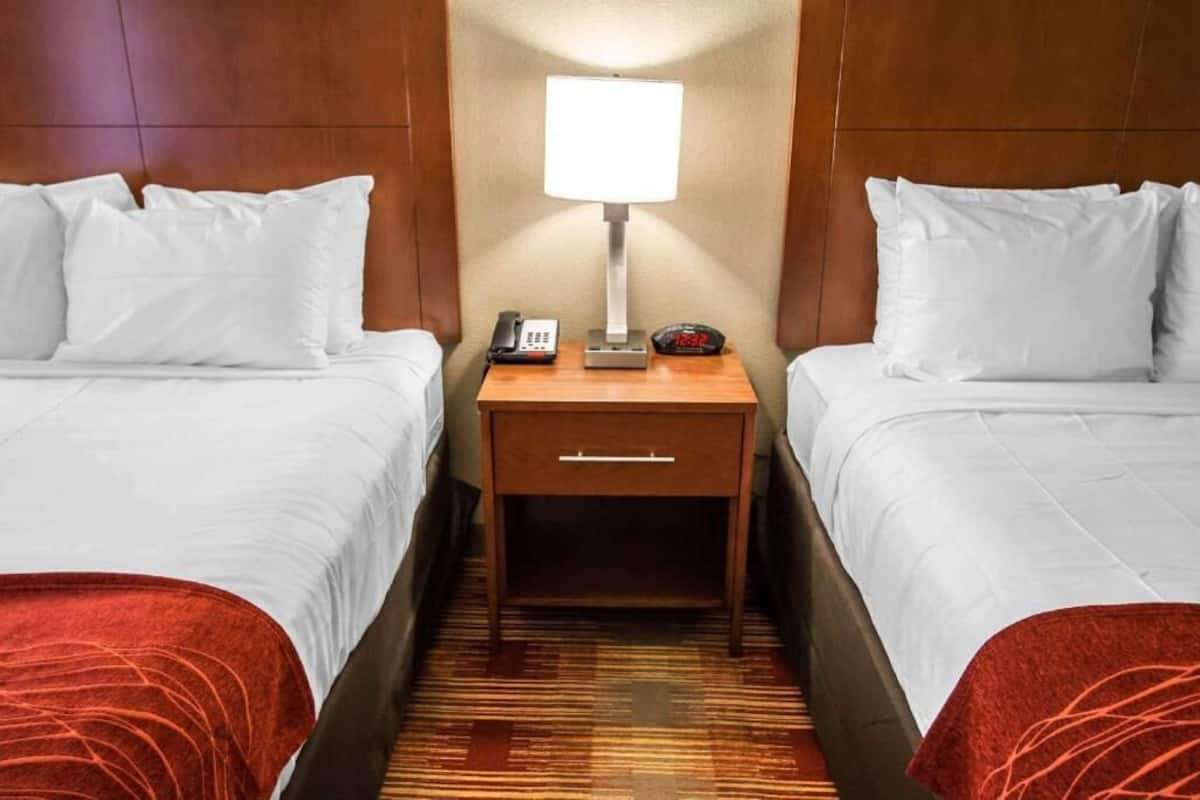 A hotel room with two beds, a nightstand with a lamp, a phone, and a clock.