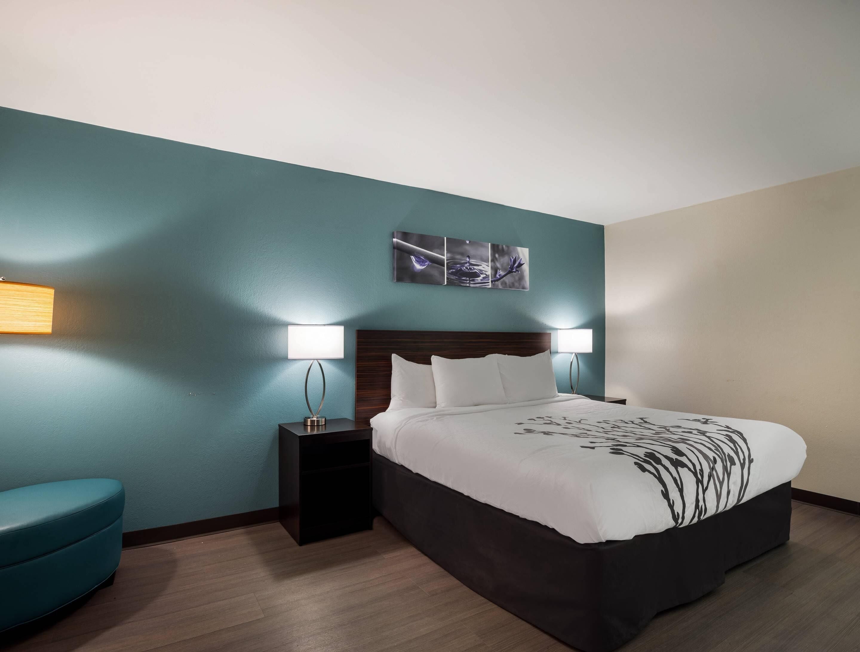 A hotel room with a bed, two bedside tables, a nightstand, a blue chair, and a wall-mounted artwork.