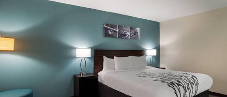 A hotel room with a bed, two bedside tables, a nightstand, a blue chair, and a wall-mounted artwork.