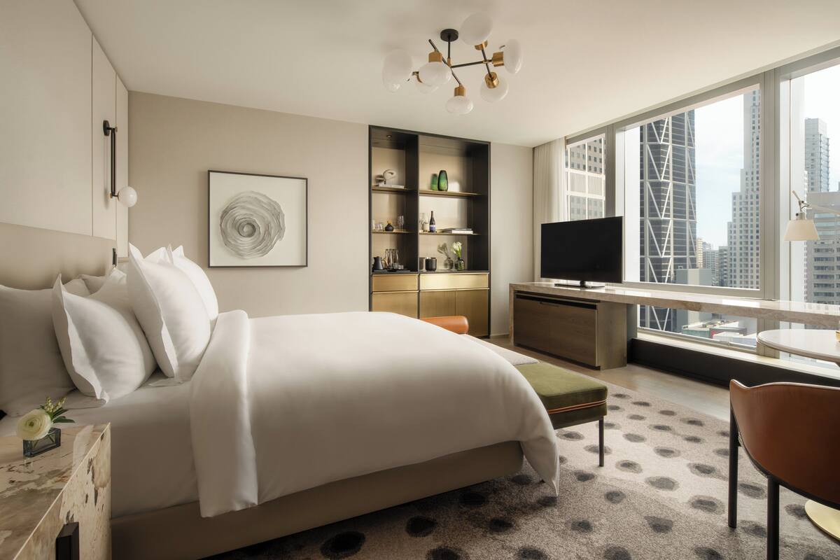 A modern hotel room with a large bed, a desk, a television, and a view of the cityscape.