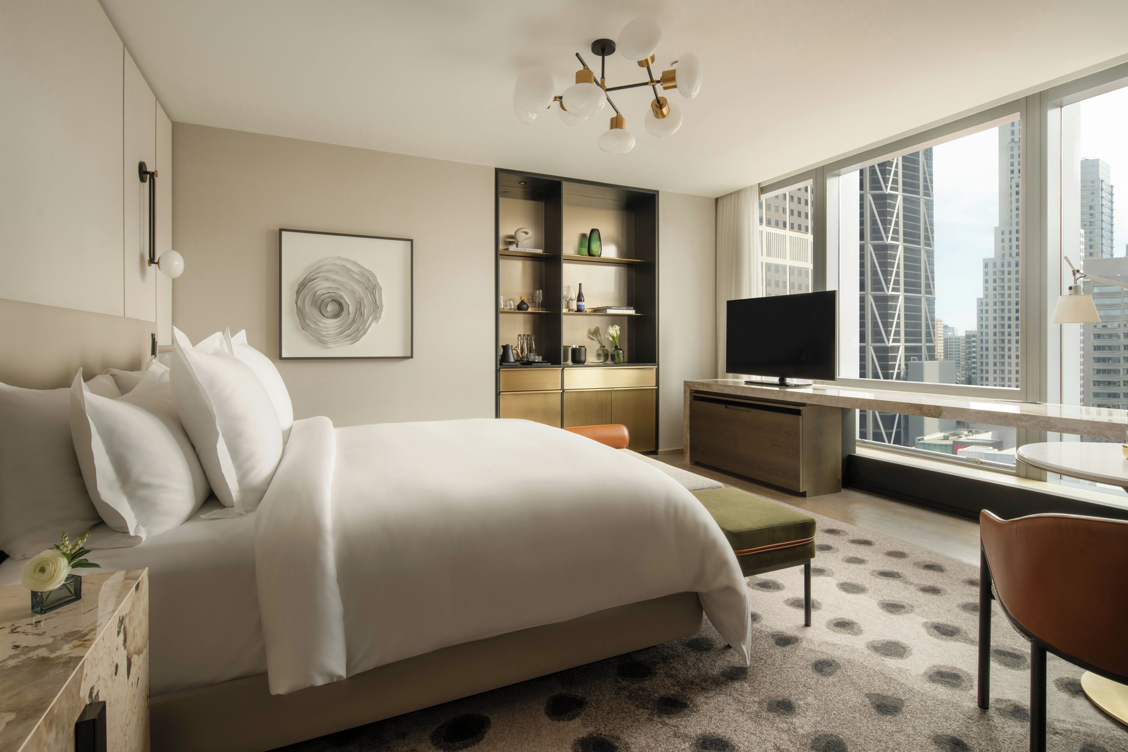 A modern hotel room with a large bed, a desk, a television, and a view of the city skyline.