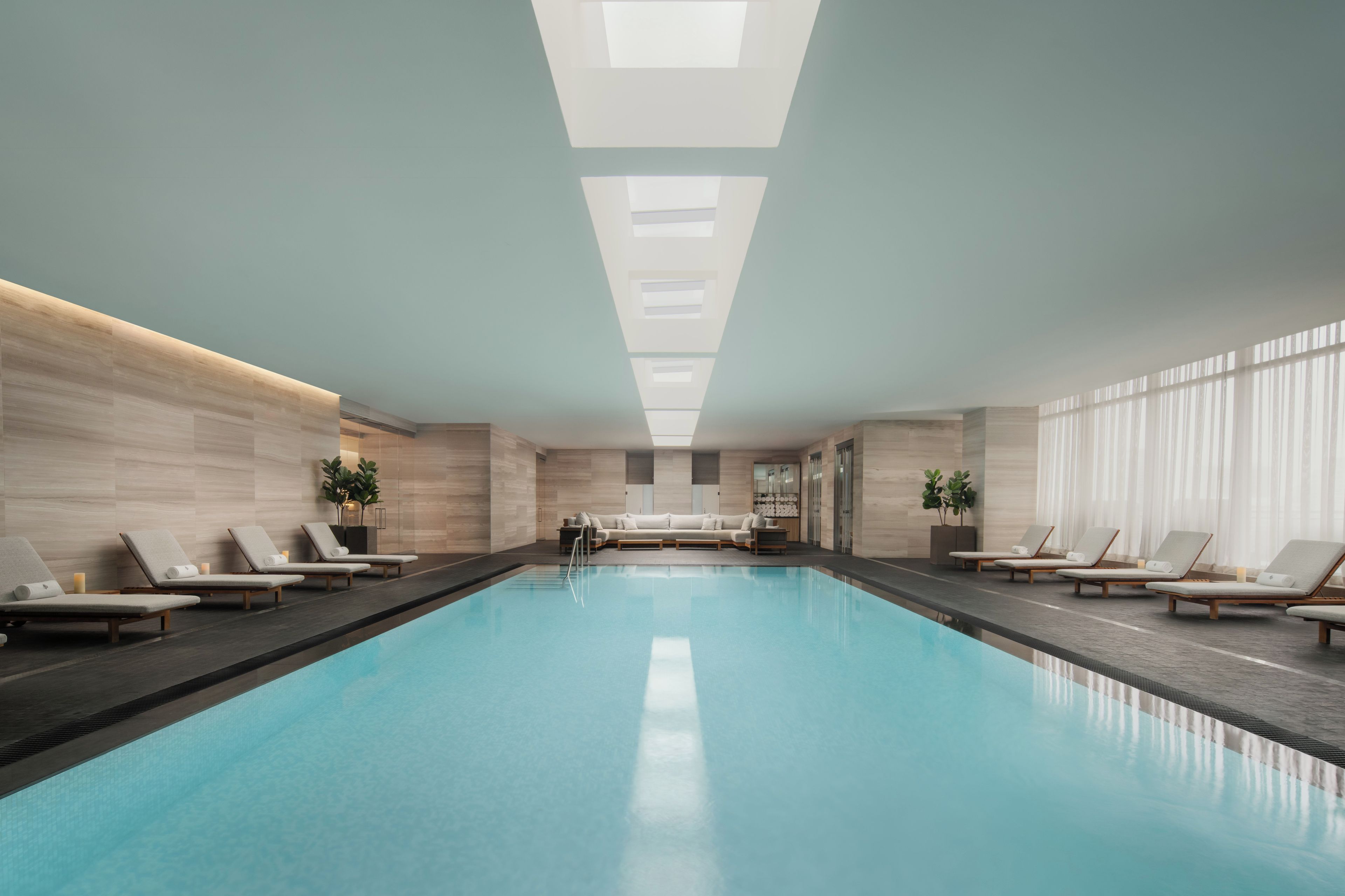 Une piscine intérieure moderne, dotée de chaises longues et d’un canapé, et entourée de murs en bois et de grandes fenêtres.