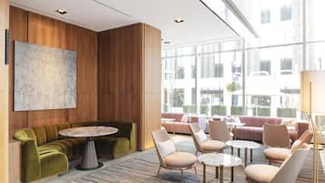 A modern lounge area with a round table, sofas, and chairs, wooden paneling, and large windows offering a view of the city.