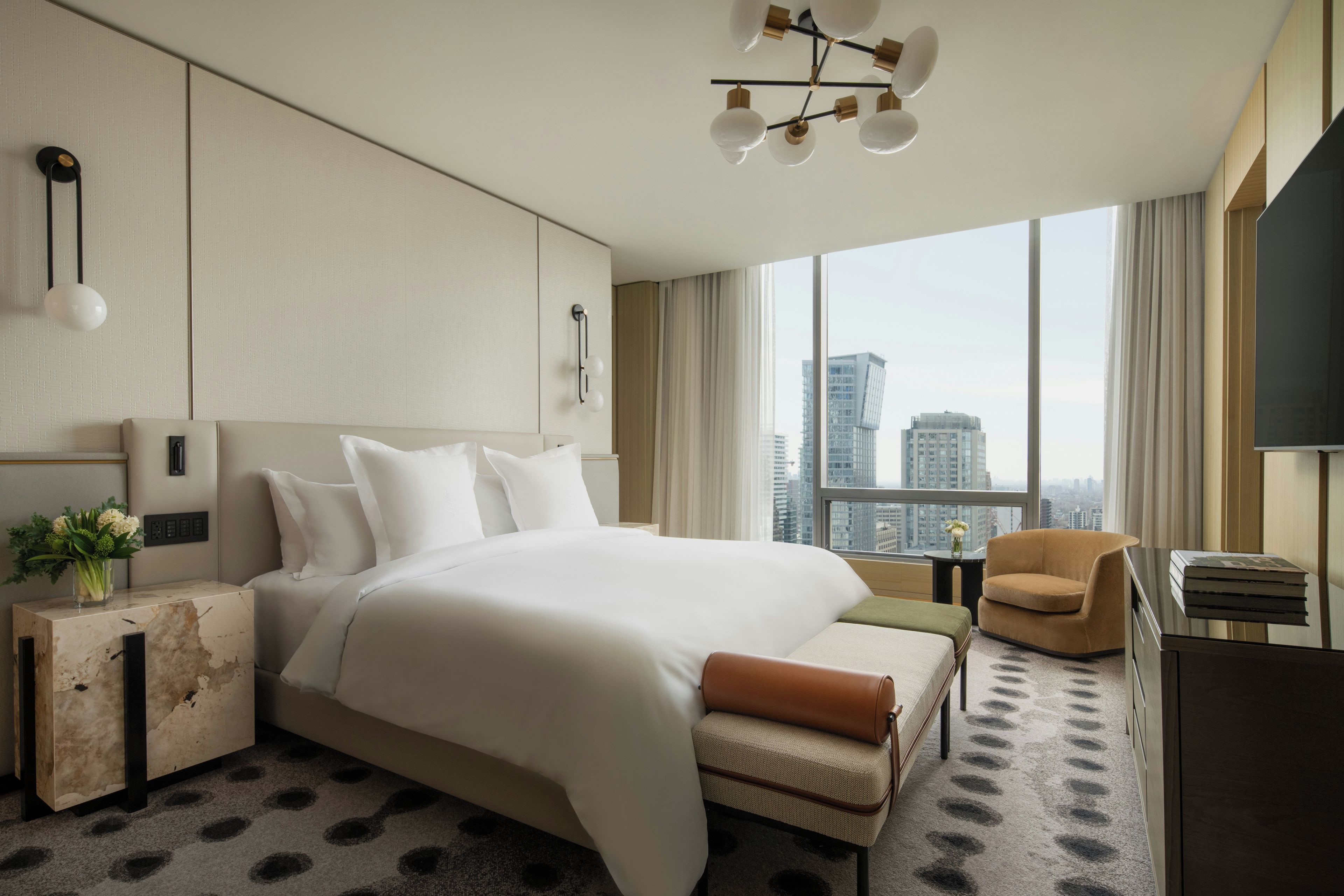 Habitación de hotel con una cama grande, un escritorio, una silla, un televisor y vista a la ciudad.