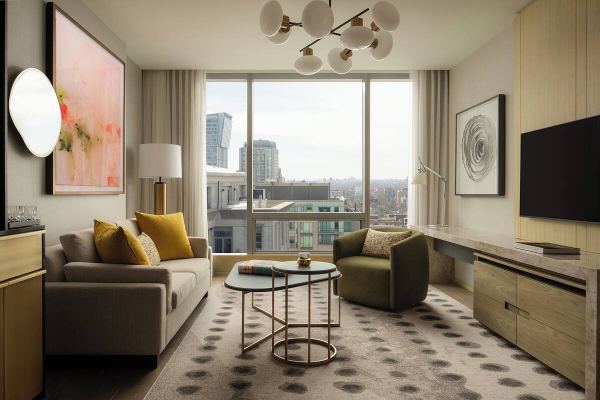 A modern hotel room with a sofa, armchair, coffee table, and large windows offering a city view.