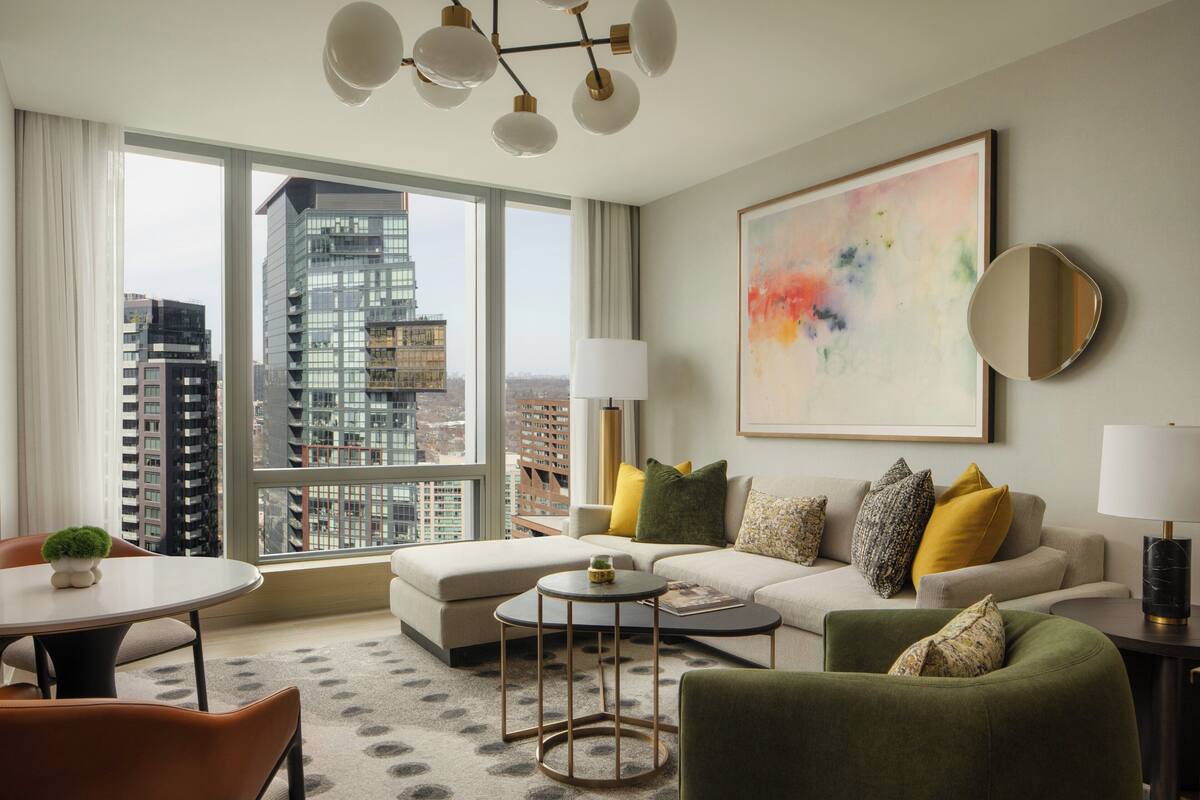 A modern living room with a sofa, armchairs, a coffee table, and a dining table. Large windows offer a view of the cityscape.