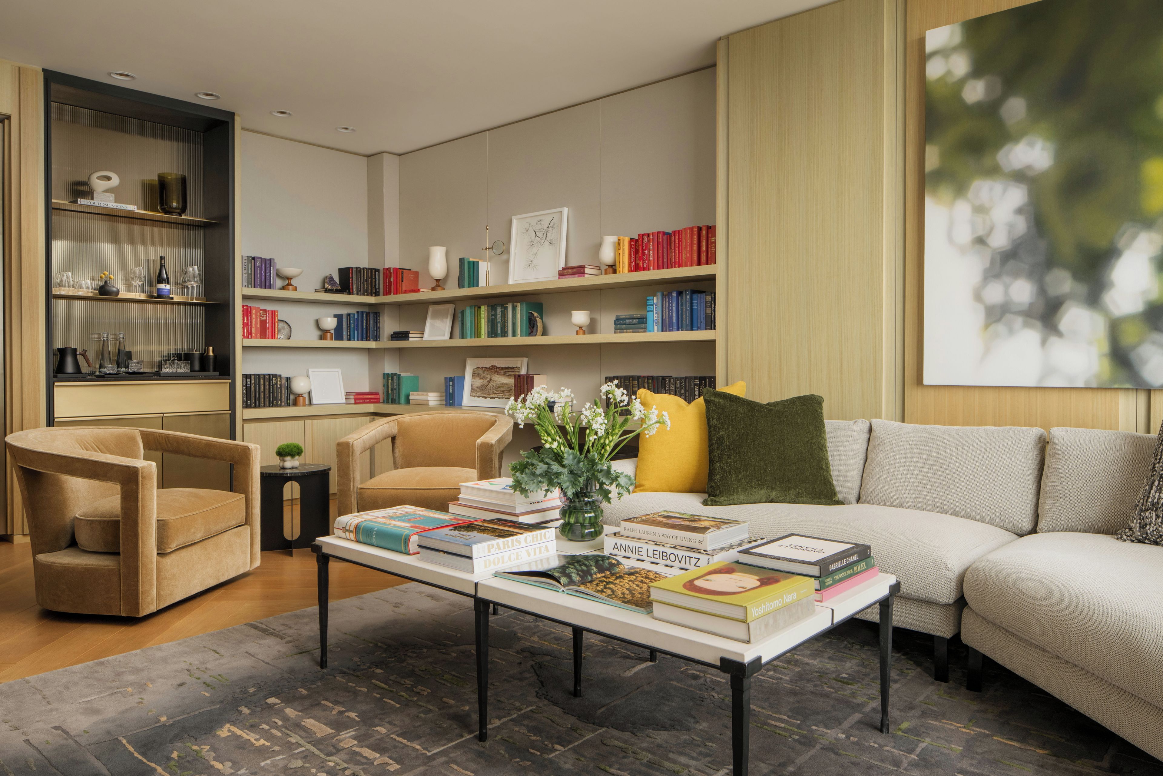 A modern living room with a sofa, armchairs, a coffee table with books, a bookshelf, and a large window with a view.