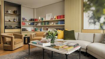 A modern living room with a sofa, armchairs, a coffee table with books, a bookshelf, and a large window with a view.