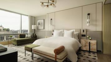 A modern hotel room with a large bed, a seating area with a sofa and armchair, a side table, and a view of the city.