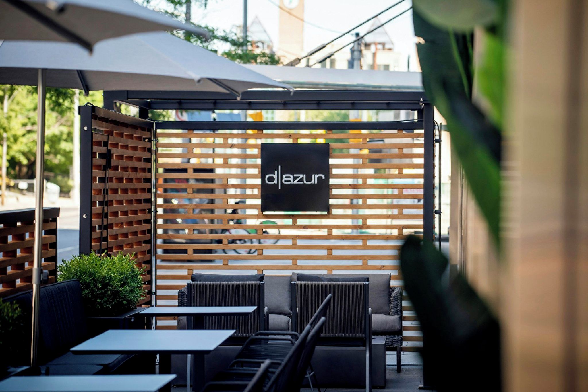 An outdoor seating area with a wooden partition, a sign reading 'd|azur', and tables and chairs.