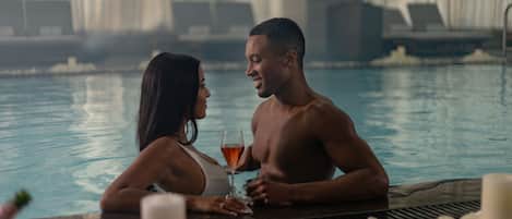 A couple in a pool with a table, candles, and a glass of drink.