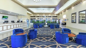 A hotel lobby with a reception desk, blue seating, a patterned carpet, and a large window with plants. - Alexis Park All Suite Resort (Las Vegas)