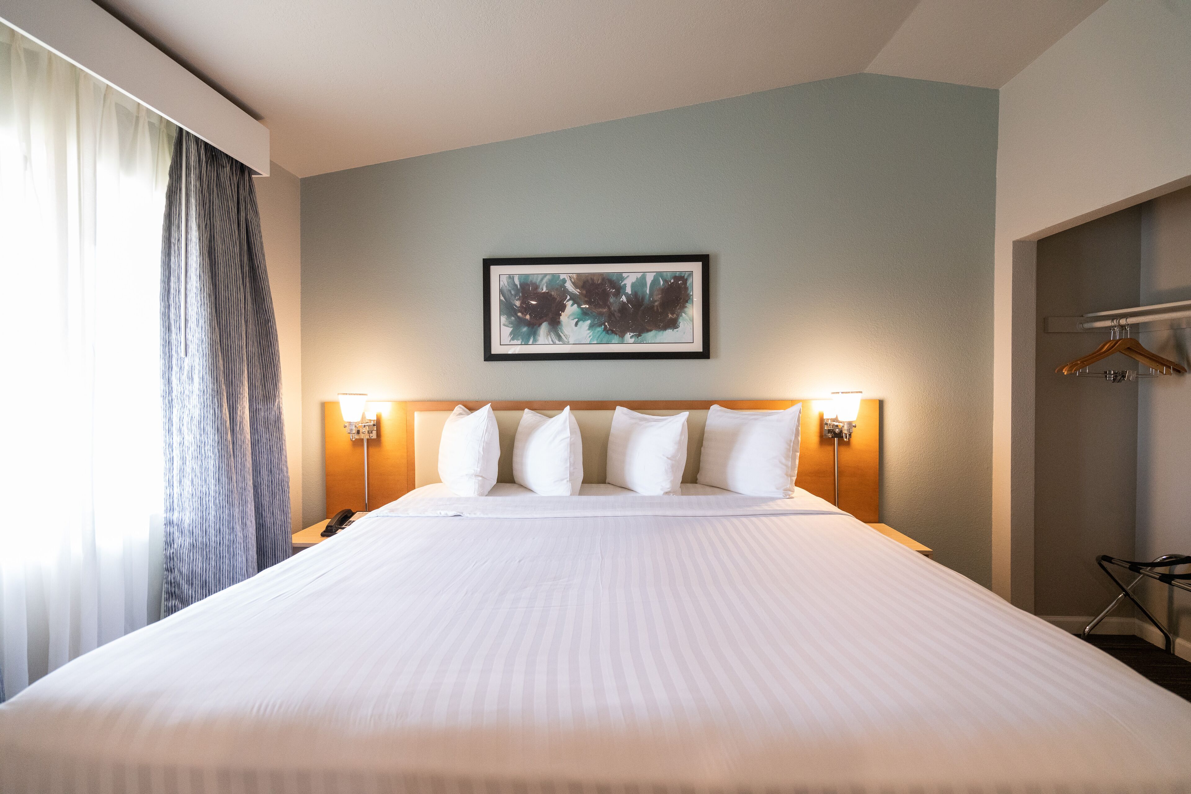 A hotel room with a large bed, a headboard with lights, a framed artwork on the wall, and a coat rack.