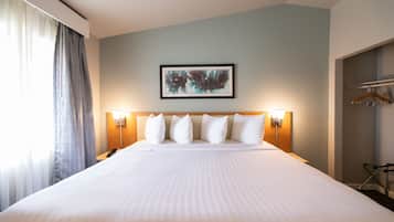 A hotel room with a large bed, a headboard with lights, a framed artwork on the wall, and a coat rack.