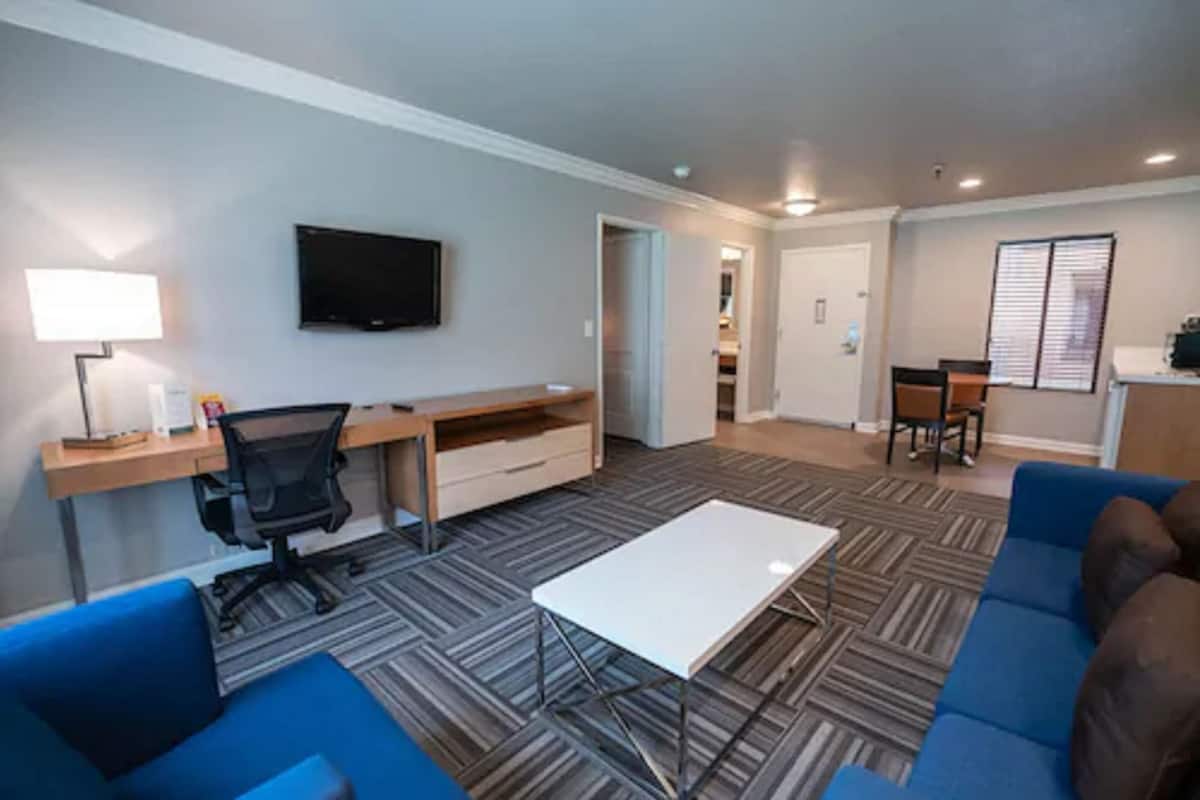 A modern living room with a flat-screen TV, a desk with a chair, a coffee table, and blue sofas.