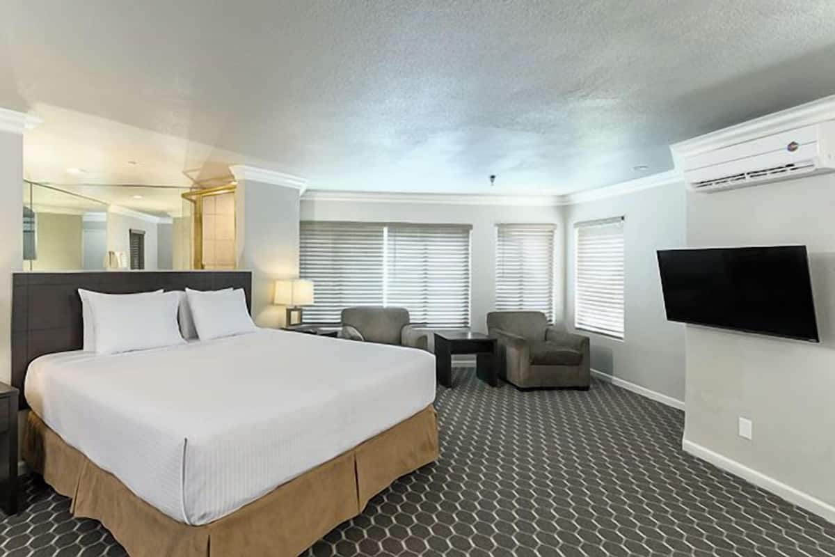 A hotel room with a large bed, a desk, two chairs, a TV, and a window with blinds.