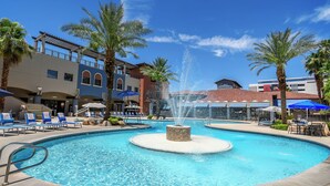 A swimming pool with a central fountain, surrounded by lounge chairs and umbrellas, with palm trees and a building in the background. - Alexis Park All Suite Resort (Las Vegas)