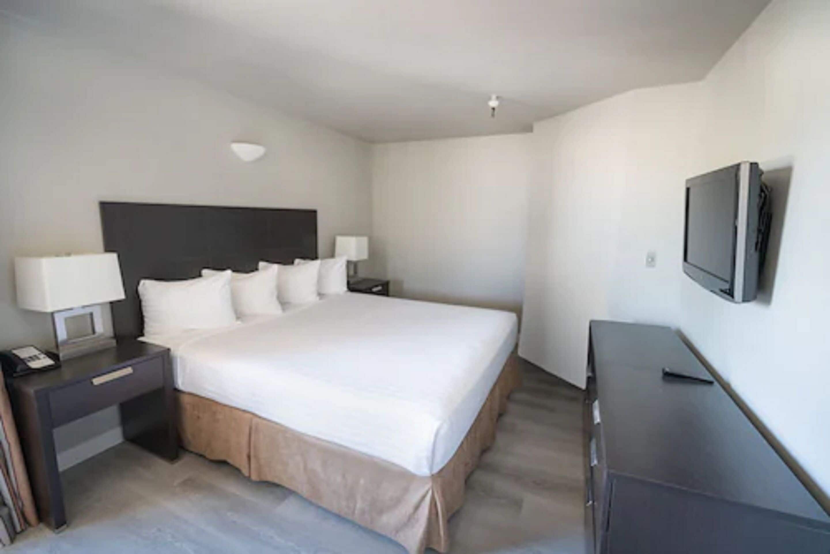 A hotel room with a large bed, a bedside table with a lamp, a TV mounted on the wall, and a dresser.