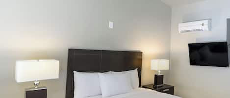 A hotel room with a large bed, two bedside tables with lamps, a flat-screen TV, an air conditioning unit, and a ceiling light.