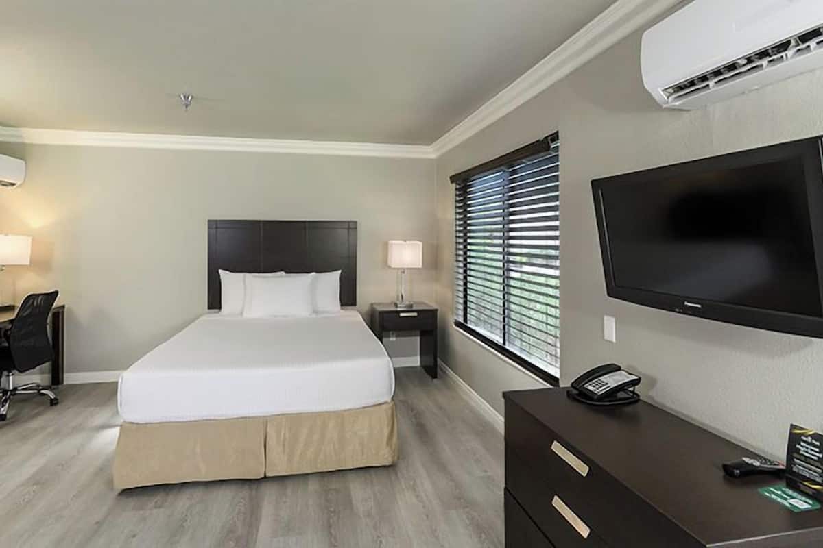 A hotel room with a bed, desk, chair, bedside tables, lamp, TV, and an air conditioning unit.