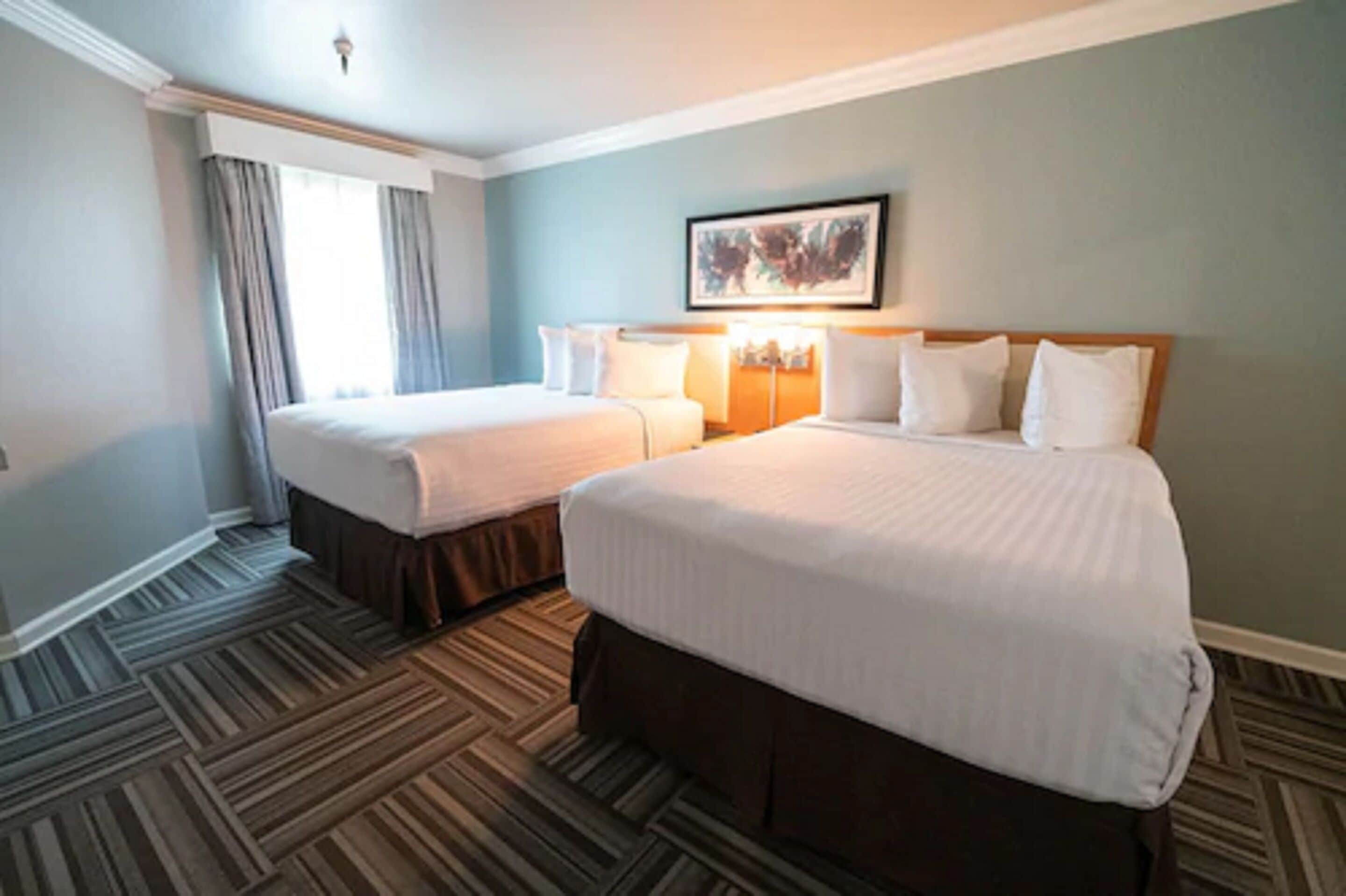 Quarto de hotel com duas camas, uma janela grande com cortinas, uma obra de arte emoldurada acima das camas e um tapete estampado.