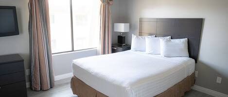Una cama bien tendida con cabecera, una mesita de noche con lámpara y una ventana con cortinas.
