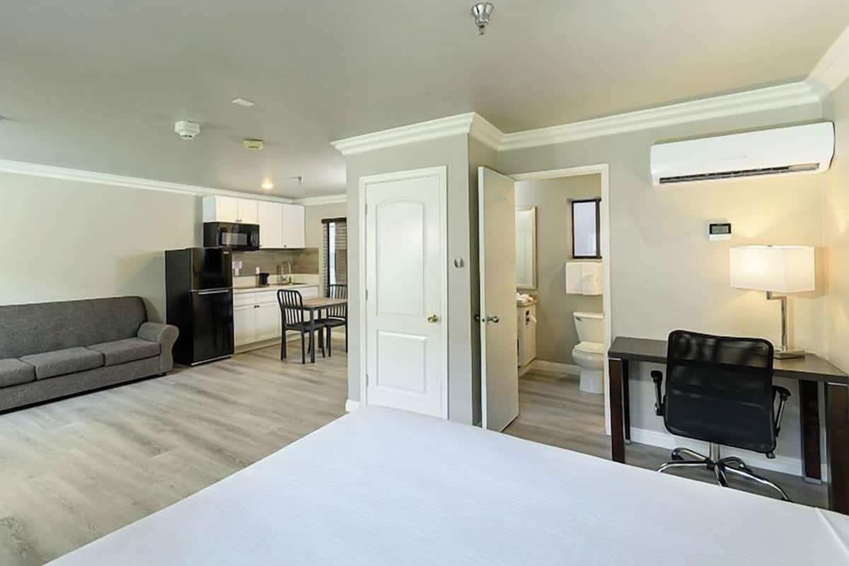 A modern hotel room with a bed, a desk, a chair, a sofa, a kitchenette, and a bathroom.