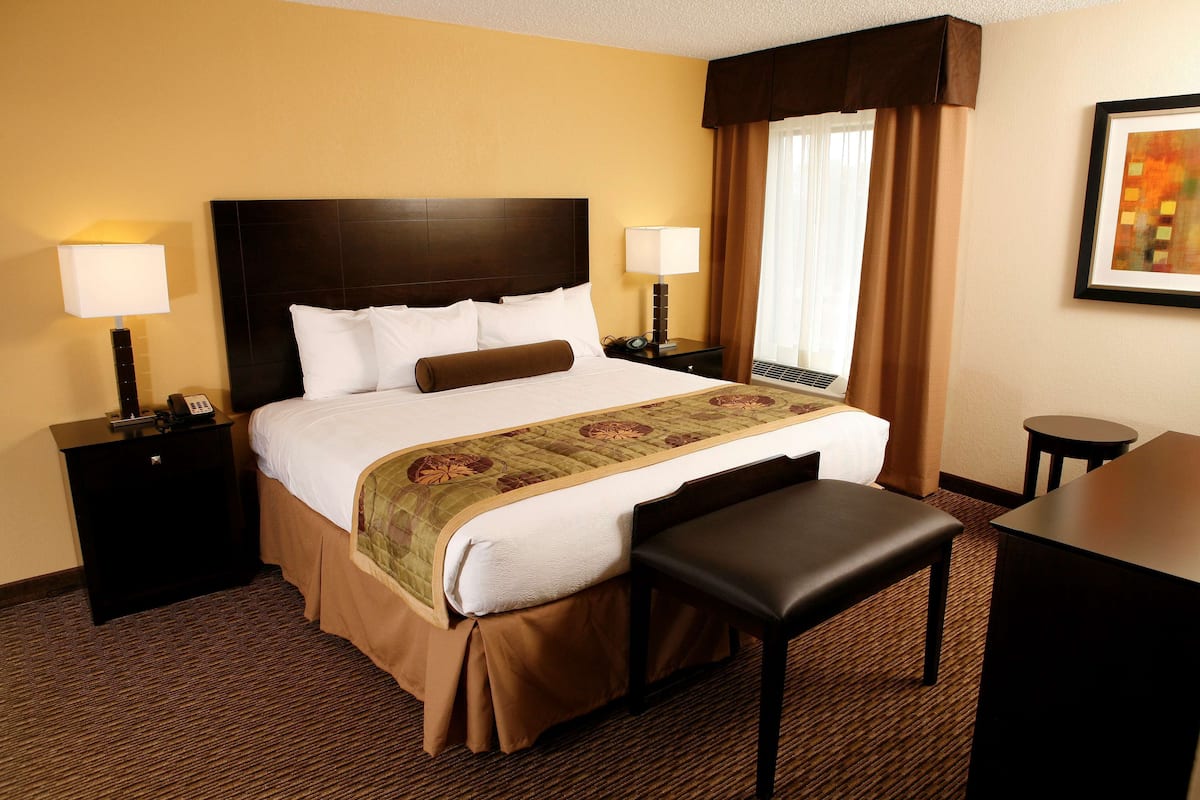 Una habitación de hotel con una cama grande, dos mesitas de noche con lámparas, un banco, una pequeña mesa y una ventana con cortinas.