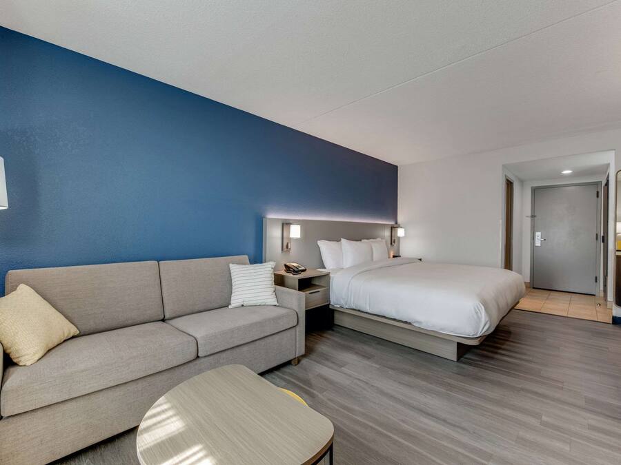 Una habitación de hotel moderna con un sofá gris, una cama, una mesita y una mesita de noche con lámpara.