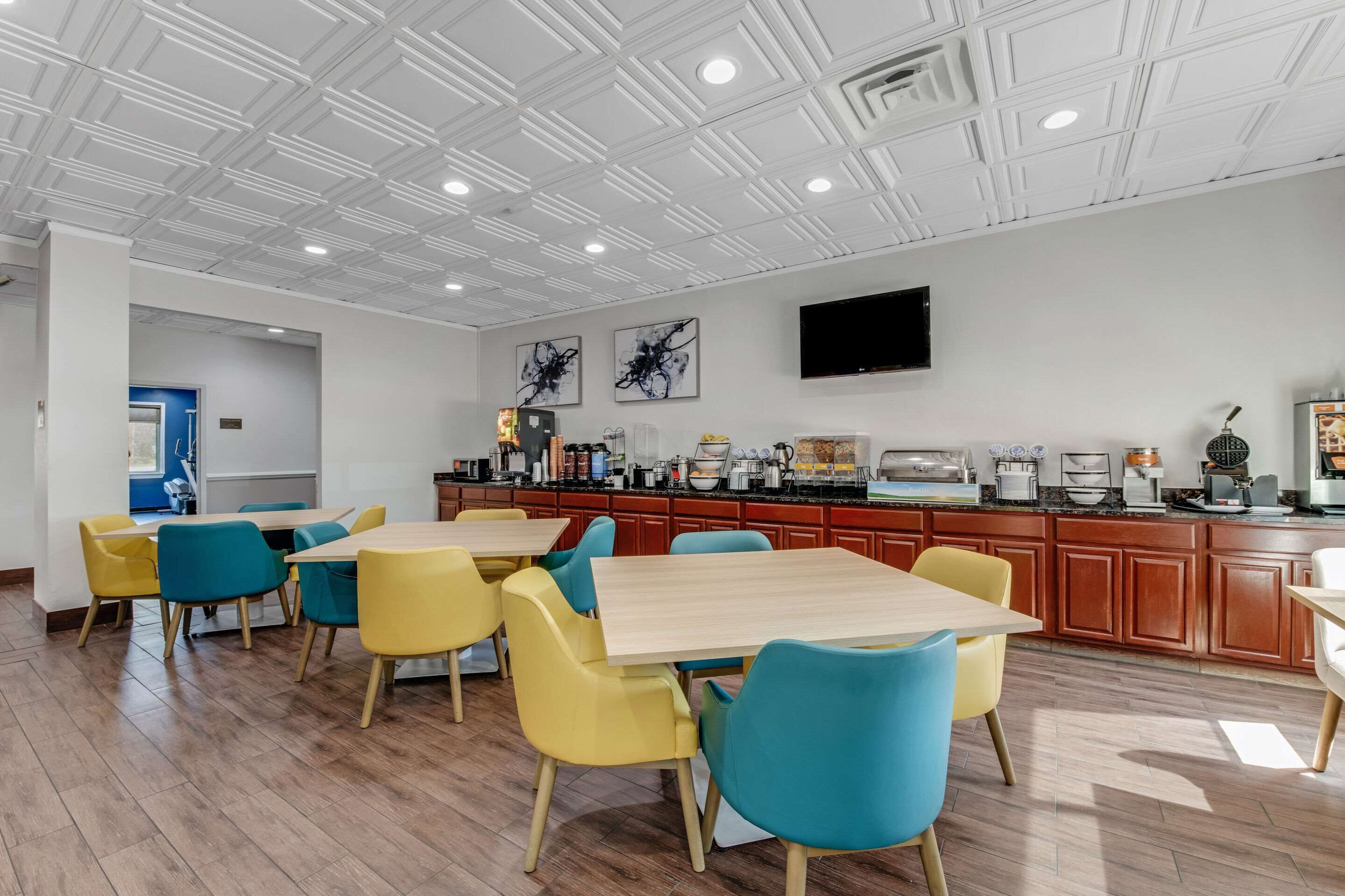 Un comedor moderno con mesas de madera y sillas coloridas, una encimera de cocina con diversos electrodomésticos y un televisor de pantalla plana montado en la pared.
