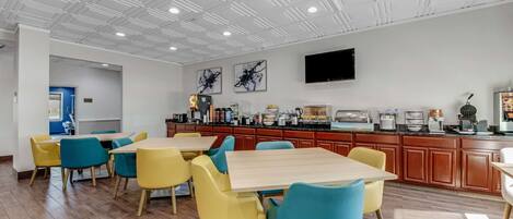 Un comedor moderno con mesas de madera y sillas coloridas, una encimera de cocina con diversos electrodomésticos y un televisor de pantalla plana montado en la pared.