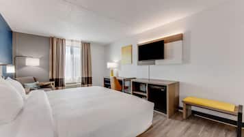 A hotel room with a large bed, a desk, a chair, a TV, and a bench.