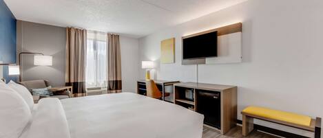 A hotel room with a large bed, a desk, a chair, a TV, and a bench.