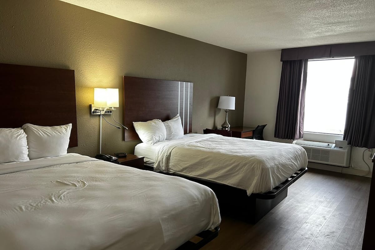 A hotel room with two beds, a desk, a chair, a lamp, and a window with curtains.