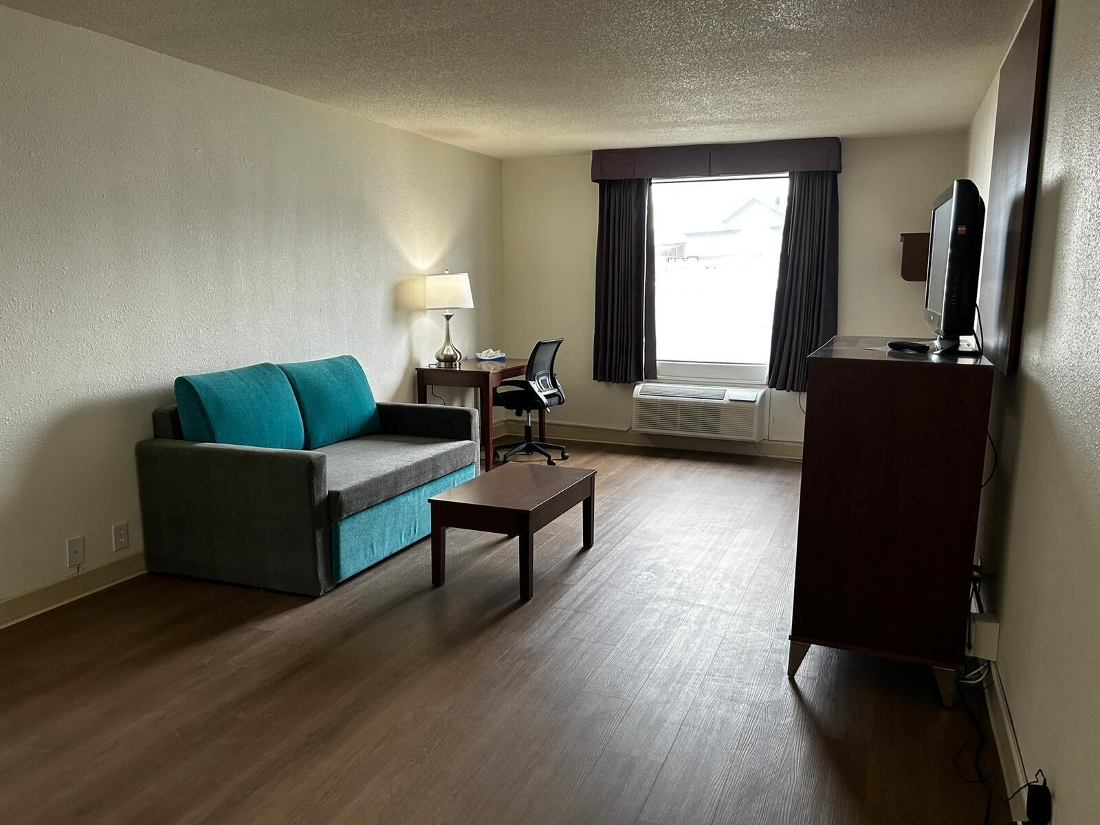 Une chambre d’hôtel équipée d’un canapé, d’une petite table, d’un bureau avec une chaise, d’un climatiseur et d’un téléviseur posé sur un meuble.