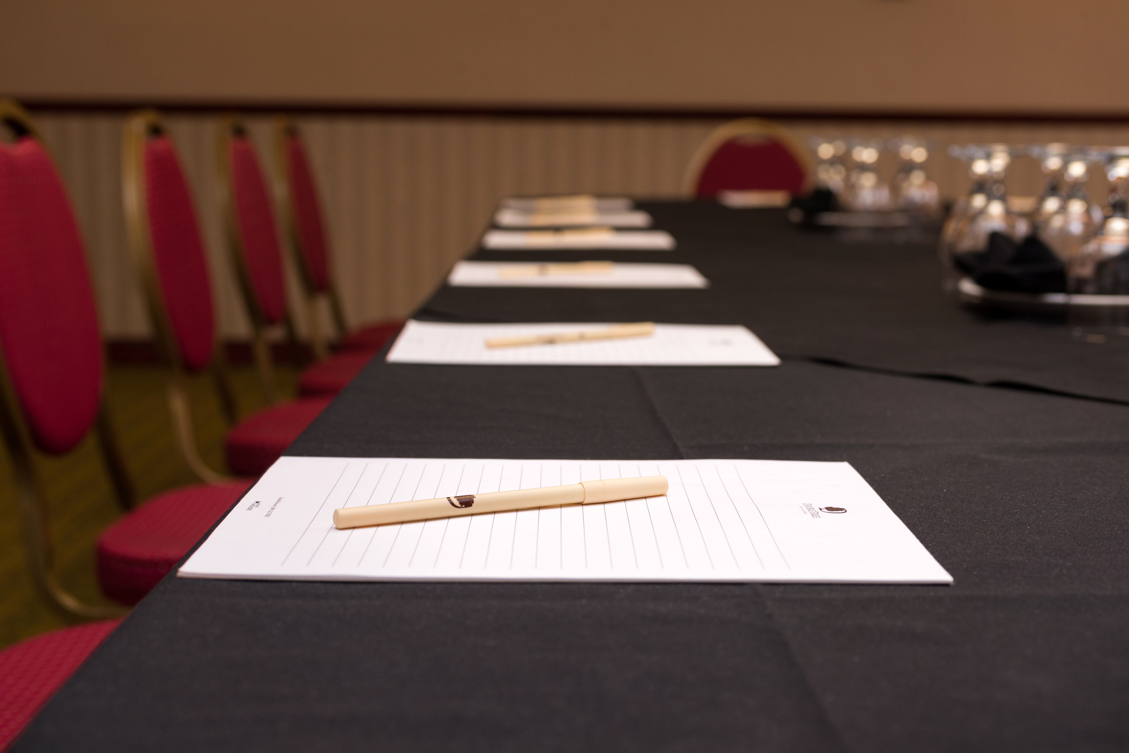 A conference table with lined sheets and a pen, set against a backdrop of red chairs and a wall with a vent.