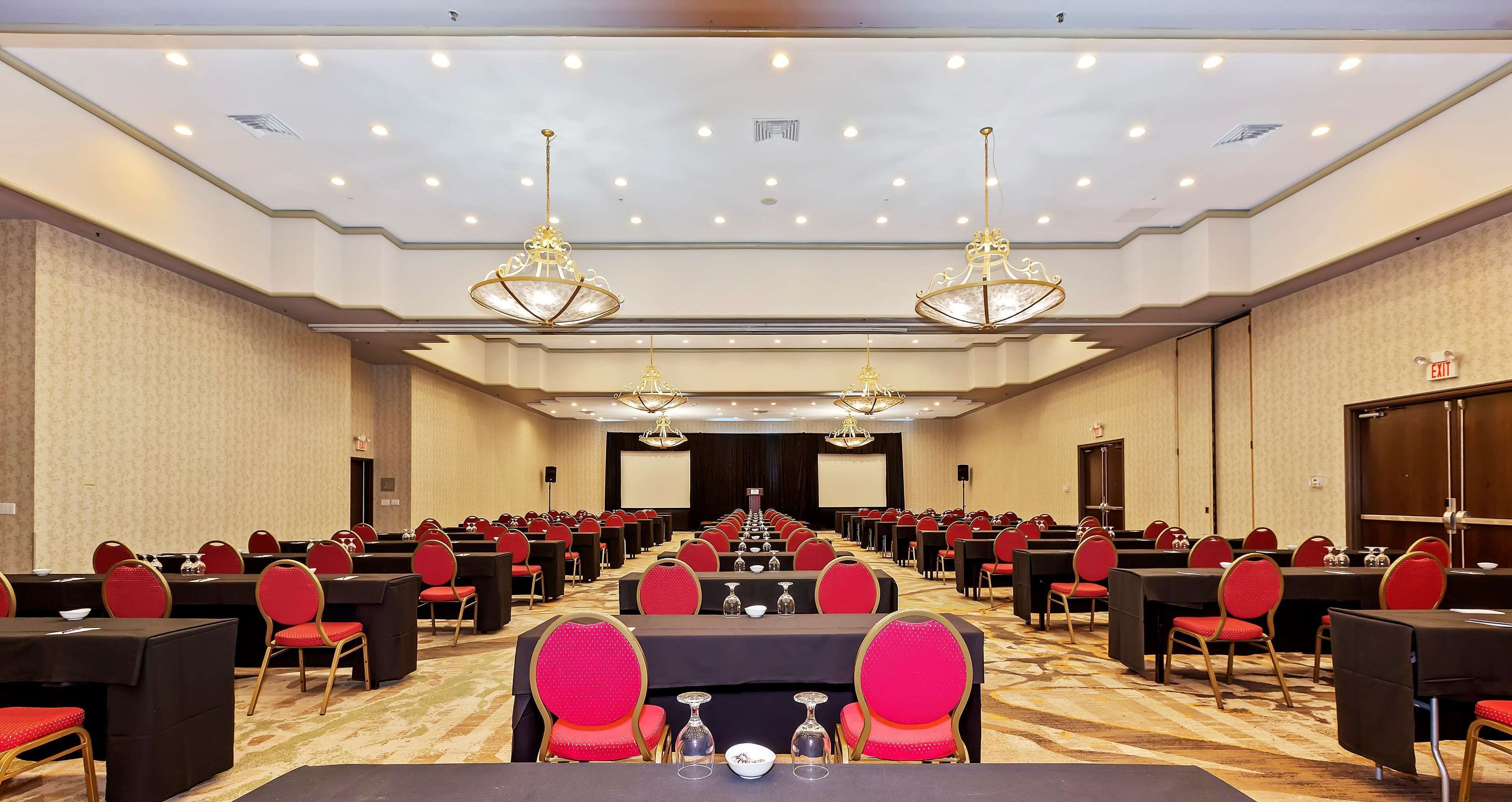 Uma sala de conferências com mesas e cadeiras dispostas em fileiras, grandes lustres e uma tela de projeção na frente.