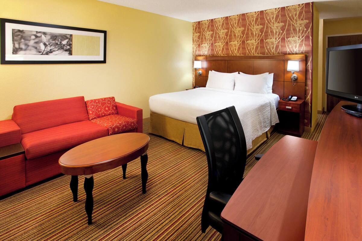 A hotel room with a bed, a red sofa, a wooden table, a TV, and a nightstand with a lamp.