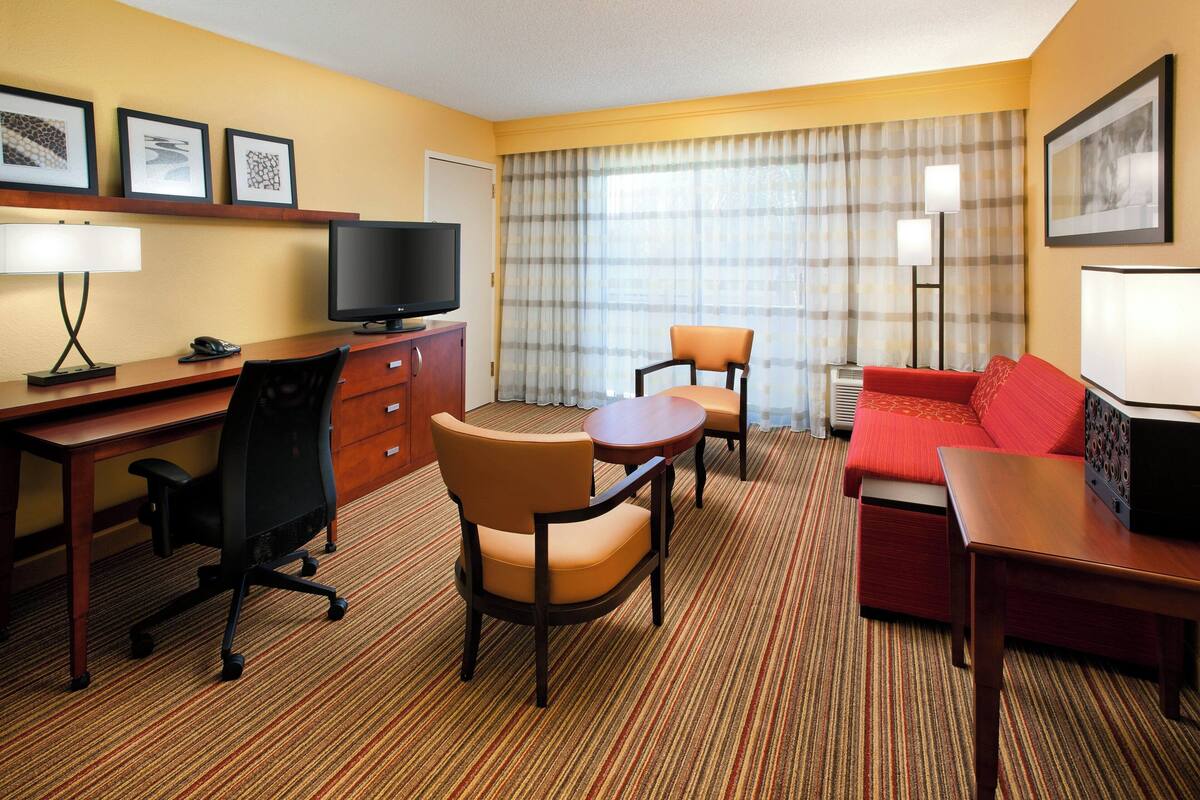 A hotel room with a desk, two chairs, a sofa, a television, and a lamp.