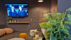 Habitación moderna con una televisión mostrando un evento deportivo, un sofá, una pequeña mesa con velas y una planta en maceta.