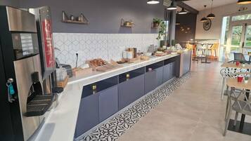 Une cuisine moderne équipée d’une machine à café intégrée, d’un comptoir où sont disposés des aliments et d’un espace repas avec des tables et des chaises.