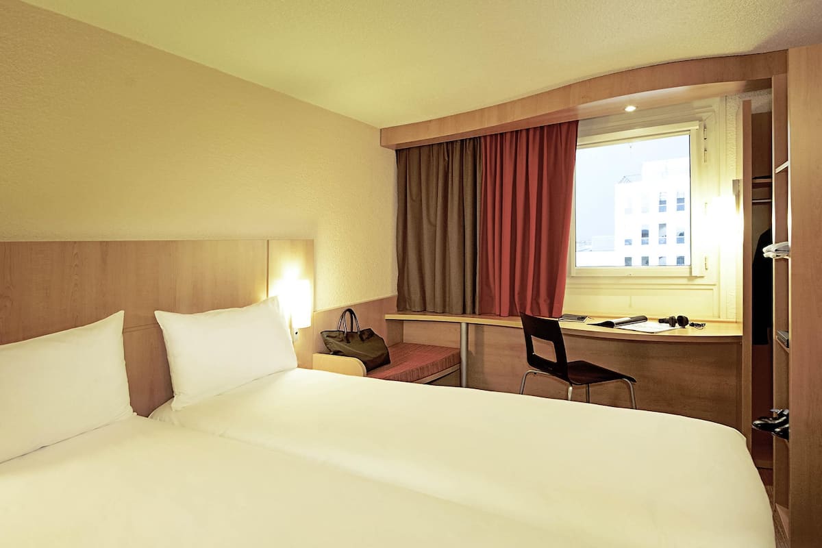 Una habitación de hotel con una cama grande, un escritorio con una silla, una ventana con cortinas y una mesita de noche con lámpara.
