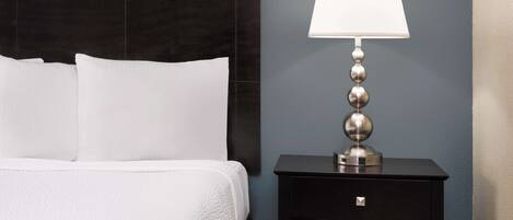 A neatly made bed with white linens and pillows, a black nightstand with a lamp, and a blue wall.