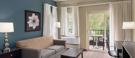 A hotel room with a sofa, a small table, a desk with a chair, and a large window with curtains.