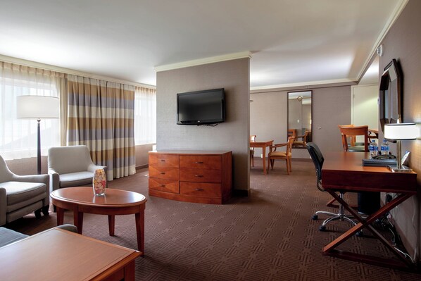 A hotel room with a flat-screen TV mounted on the wall, a wooden dresser, a round table with a vase of flowers, a desk with a chair, and a large mirror.